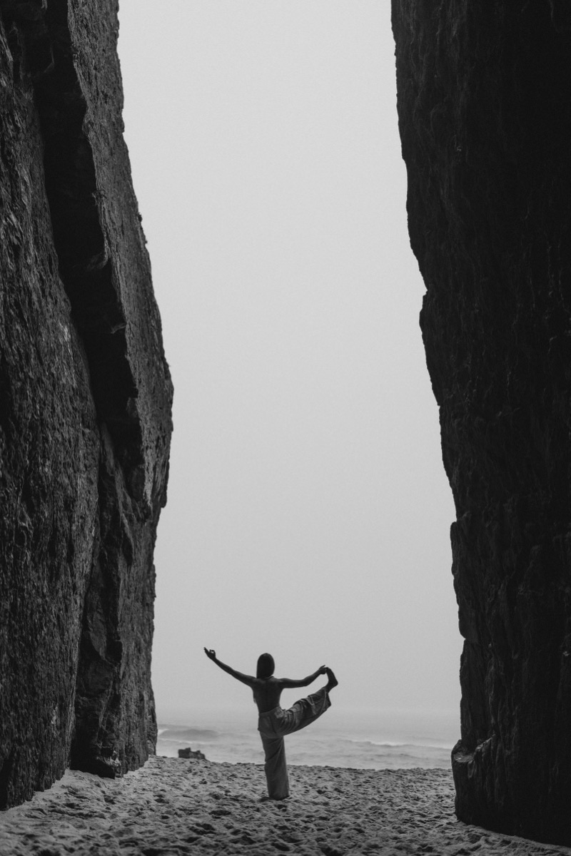 Woman in a standing balance yoga pose between rock formations on a misty beach, black and white