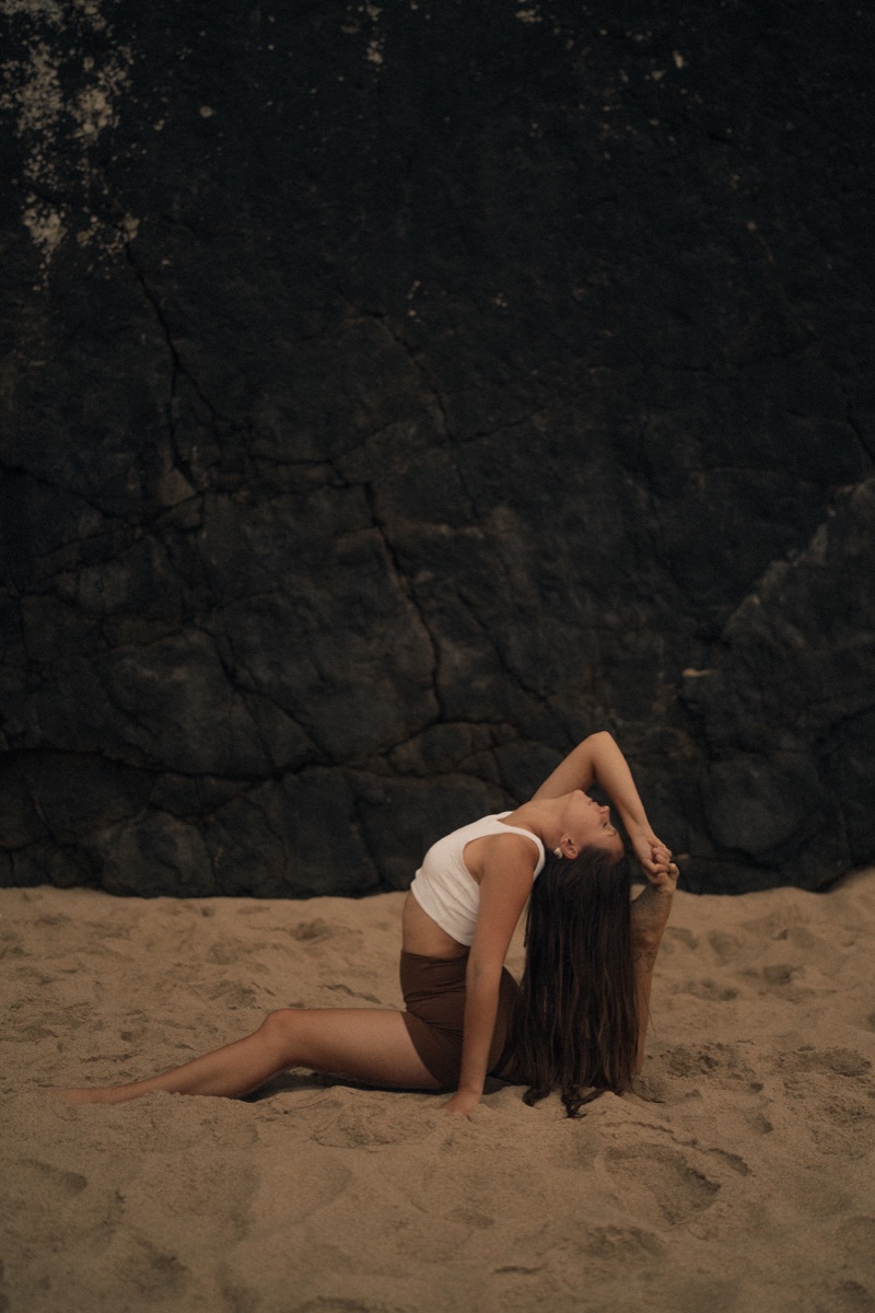 Woman in an advanced yoga pose on sand against a dark rock wall